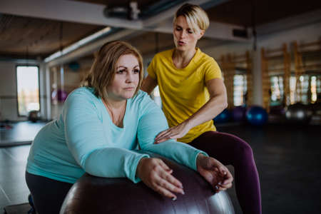 Happy overweight woman exercising with personal trainer on fintess ball in gymの写真素材