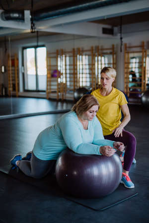 Overweight woman exercising with personal trainer on fintess ball in gymの写真素材
