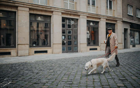 Side view of elegant senior man with take away coffee walking his dog outdoors in city in winter.