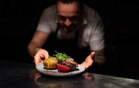Chef handing plate with meal through order station in the commercial kitchen on black background.