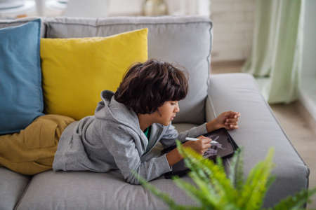 Little multiracial boy with tablet lying on sofa at home.の写真素材