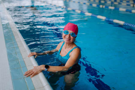 Happy senior woman in swimming pool, leaning on edge and looking at camera.