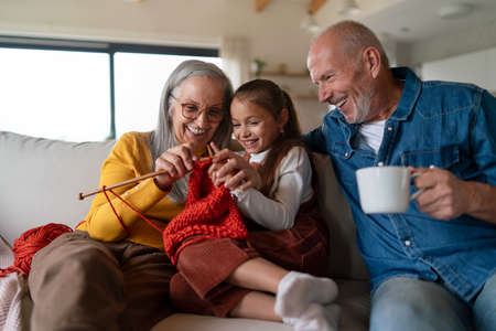 Little girl sitting on sofa with her grandparents and learning to knit indoors at home.の写真素材