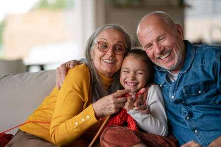 Little girl sitting on sofa with her grandparents and learning to knit indoors at home.の写真素材
