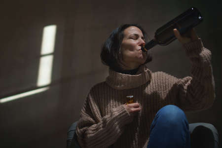 Depressed drunk woman sitting on the floor in the dark taking pills and drinking wine from a bottleの写真素材