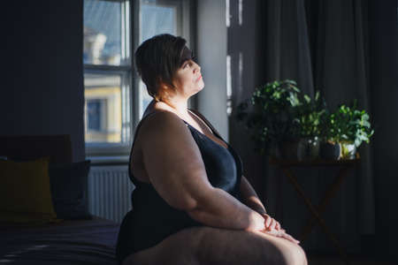 Depressed lonely fat woman sitting and looking through the window at home.の写真素材