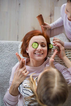 Two little girls putting cucumber on their mothers face and combing her hair at home.の写真素材