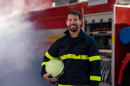 Happy mid-adult firefighter man looking at camera with fire truck and smoke in background.