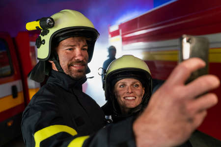 Happy firefighters man and woman after action taking selfie with fire truck in background at nightの写真素材