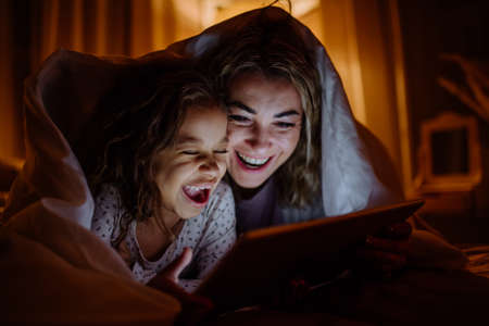Happy mother with her little daughter lying under blanket and watching movie on tablet at home.の写真素材