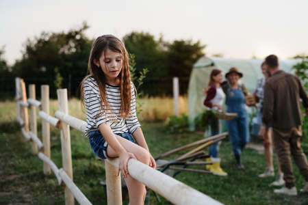 Little girl sitting on fence outdoors with her family at background at community farm.の写真素材
