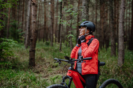 Senior woman biker putting on cycling helmet outdoors in forest in autumn day.