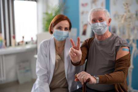 Senior man showing victory sign after being vaccinated in doctors office, looking at camera.の写真素材