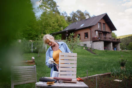 Senior woman with paintbrush impregnating wooden crate outdoors in garden.の写真素材