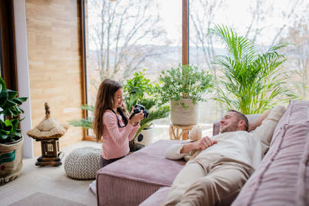 Little girl taking picture with camera of her father lying on sofa at home.