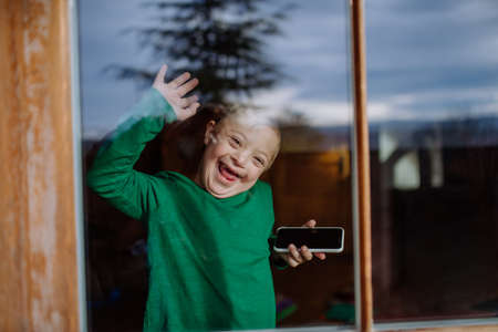 Happy little boy with Down syndrome using smartphone and waving through windowの写真素材