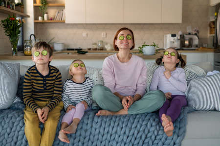 Three little children with mother sitting on sofa and putting cucumber on faces at home.