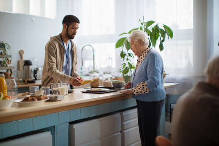 Young caregiver serving breakfast to elderly woman in nursing home care center.