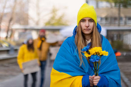 Protest against Russian invasion of Ukraine. Young woman wrapped in Ukrainian flag holding blue and yellow flowers.の写真素材