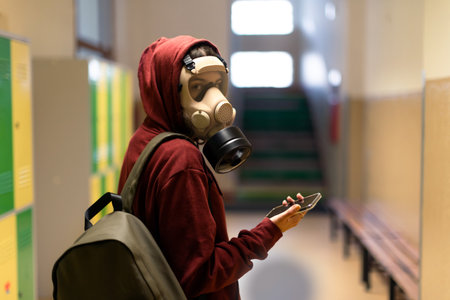 Young student in gas mask with smartphone at school.