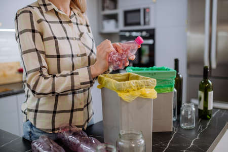 Midsection of woman throwing empty plastic bottle in recycling bin in kitchen.