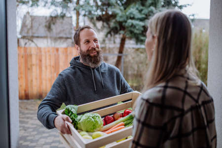 Mature man holding crate with vegetales and fruit and delivering it to woman standing at doorway.の写真素材