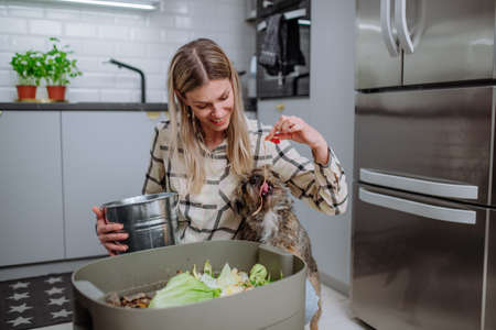 Woman throwing vegetable cuttings in a compost bucket in kitchen and feeding dog.