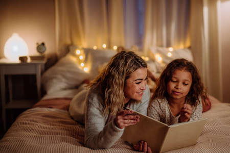 Happy mother with her little daughter lying on bed and reading book in evening at home.の写真素材