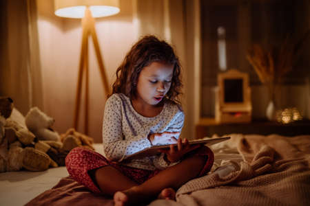 Happy little multiracial girl using tablet on bed in evening at home.