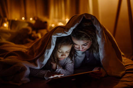 Happy mother with her little daughter lying under blanket and watching movie on tablet at home.の写真素材