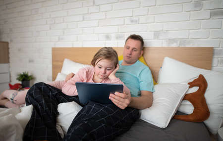 Father and his little daughter with Down syndrome lying on bed and using tablet at home.の写真素材