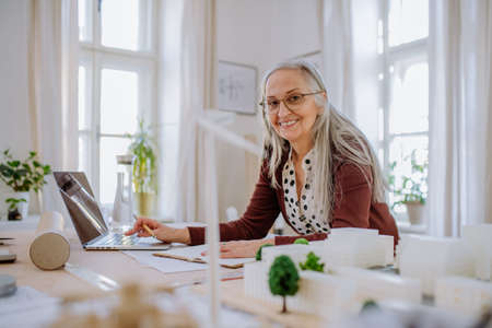 Senior woman architect with model of houses working on laptop in office and looking at camera.