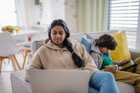 Indian mother using laptop and her little son using tablet and sitting on sofa at home.