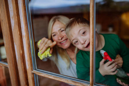 Boy with Down syndrome with his grandmother cleaning window at home.の写真素材