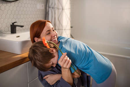 Little boy congratulates mother and gives her flower in bathroom at home.