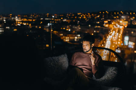 Young African AMerican man sitting on balcony with urban view and using tablet at nightの写真素材