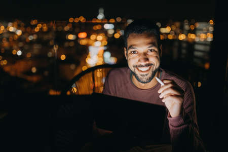 Young African AMerican man sitting on balcony with urban view and using tablet at nightの写真素材