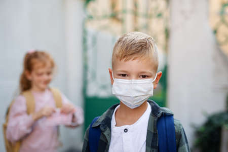 Happy little schoolboy with face mask looking at camera in front of school.
