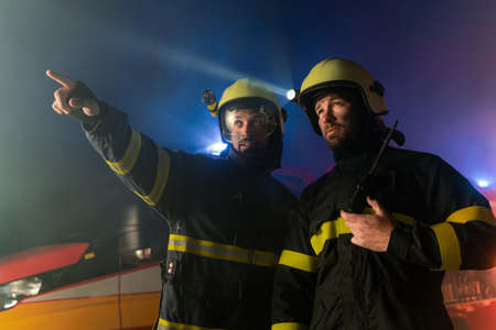 Firefighters men at action with fire truck in background at nightの写真素材