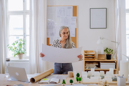 Senior woman architect with model of houses looking at blueprints in office.の写真素材
