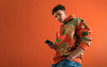 Fashion studio portrait of a young man in hoodie with smartphone posing over orange background.の写真素材