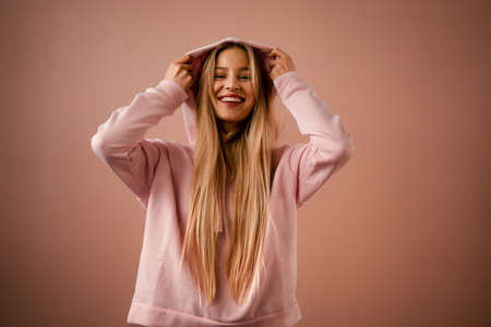 Fashion studio portrait of a happy young blonde woman in hoodie posing over pink background.の写真素材