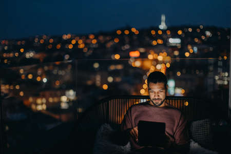 Young African American man sitting on balcony with urban view and using tablet at nightの写真素材