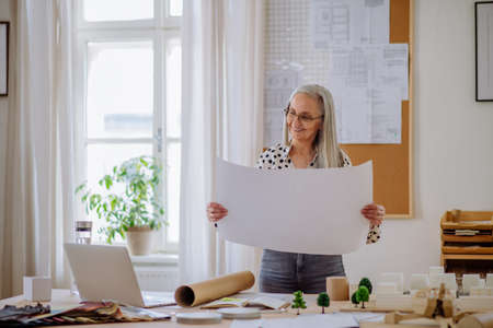 Senior woman architect with model of houses looking at blueprints in office.