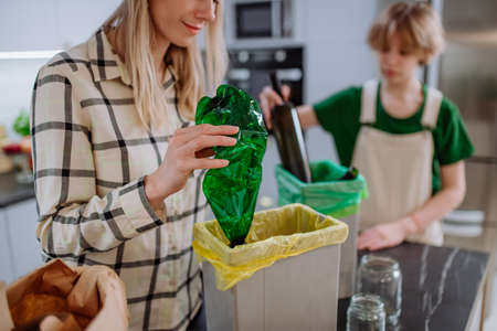 Mother with daughter throwing empty plastic and glass bottles in recycling bin in kitchen.の写真素材