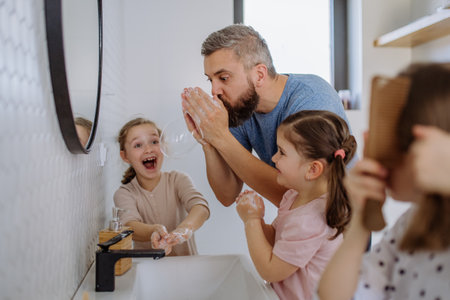 Father having fun blowing bubbles from soap in bathroom with his little daughters.