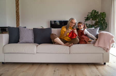 Little girl sitting on sofa with her grandmother and learning to knit indoors at home.の写真素材