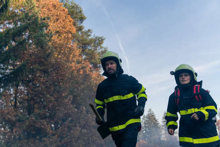 Firefighters man and woman at action, running through smoke with shovels to stop fire in forest.