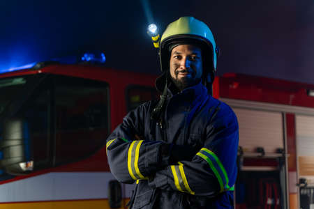 Portrait of dirty firefighter man on duty with fire truck in background at night, smiling.