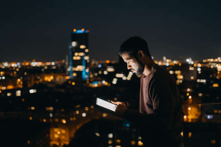 Young African American man standing on balcony with urban view and using tablet at night